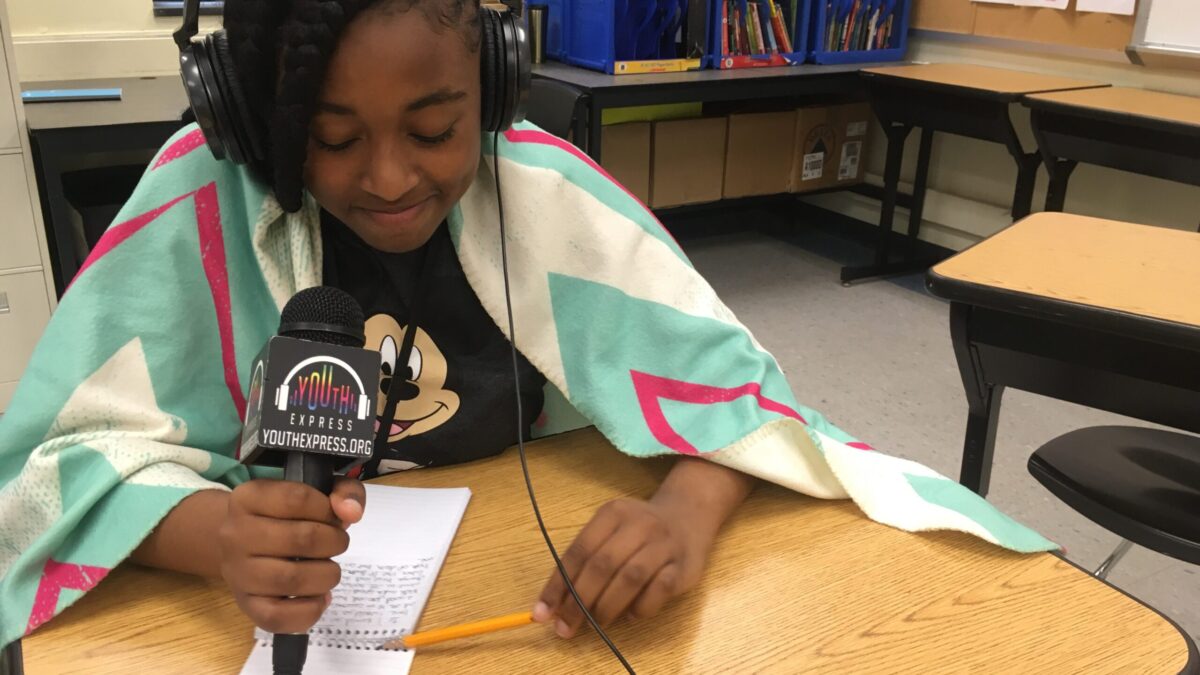 A teen looking down at a notepad and holding a pencil and a microphone sitting behind a wooden desk in a classroom