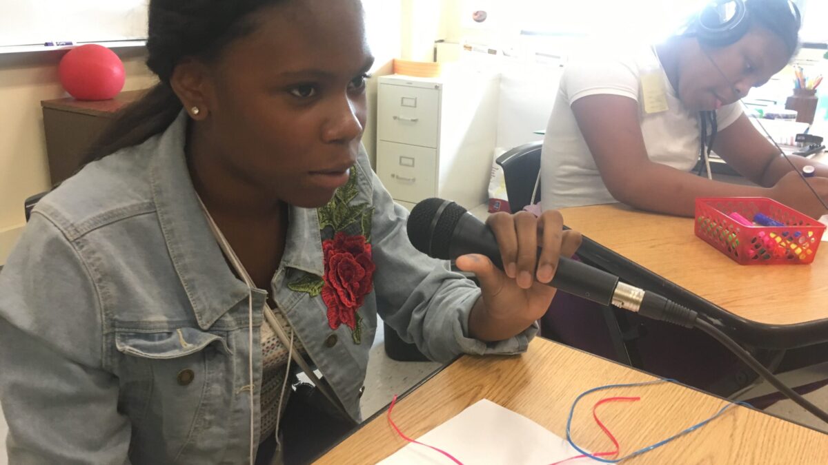 A teen speaking into a microphone and other teen wearing headphones drawing with a marker, both sitting at wooden desks in a classroom