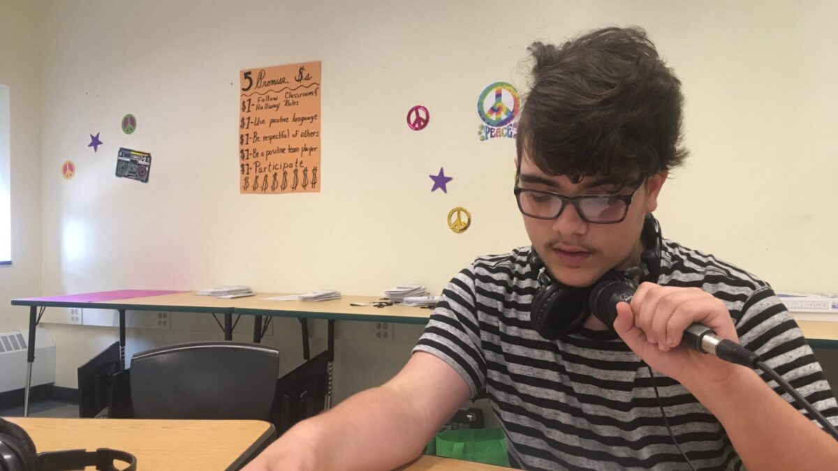 A teen looking down at a piece of paper on a wooden desk and speaking into a microphone in a classroom