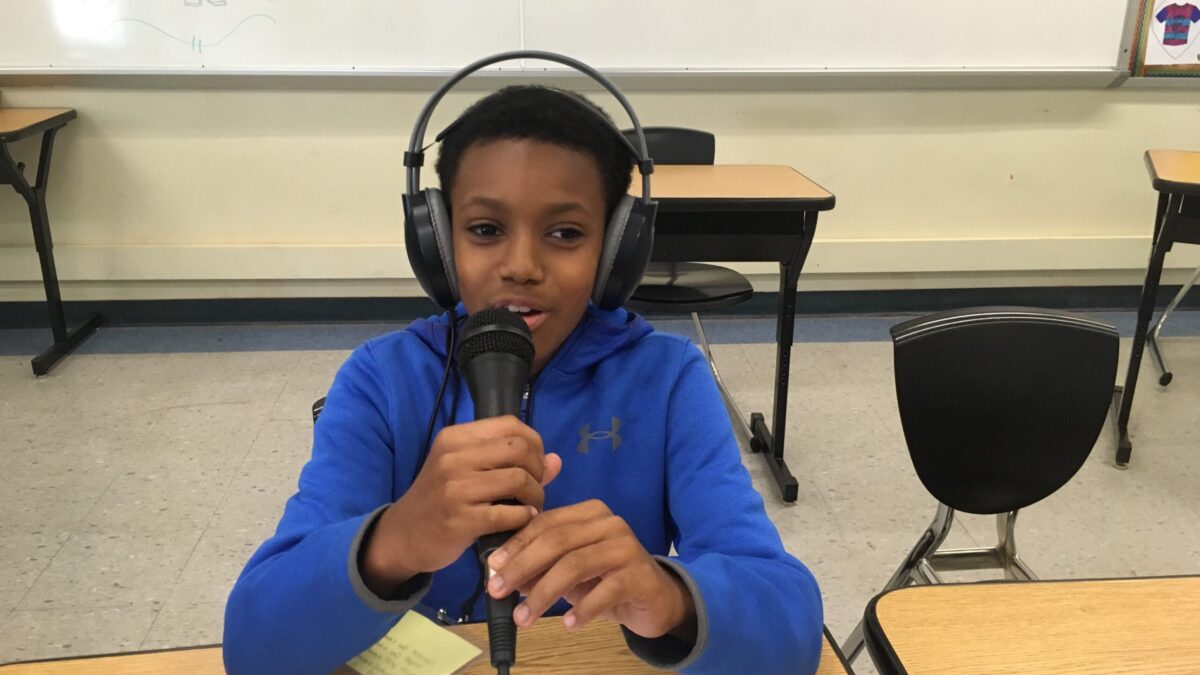 A youth wearing headphones speaking into a microphone sitting behind a wooden desk in a classroom