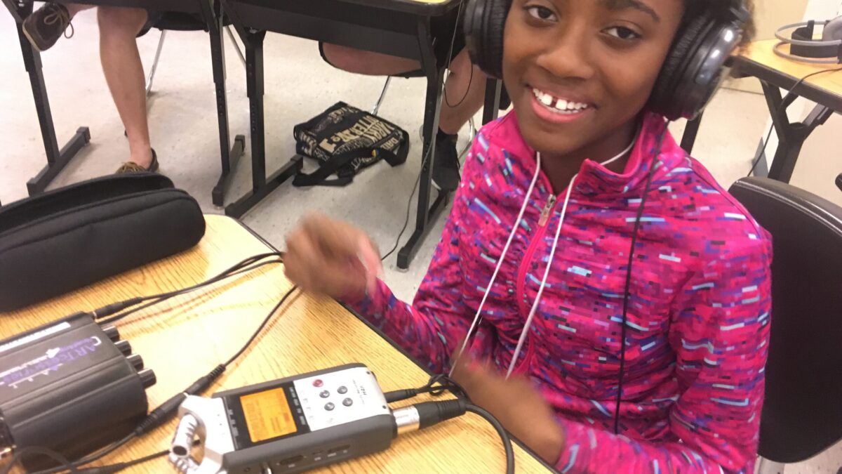 A youth wearing headphones smiling while listening to a portable recorder on a wooden desk in a classroom