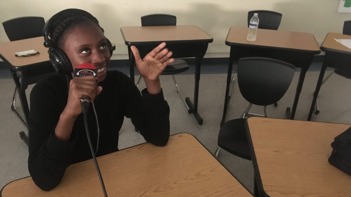 Teen wearing headphones and speaking into a microphone while gesturing to the ceiling with their left hand sitting behind a wooden desk in a classroom