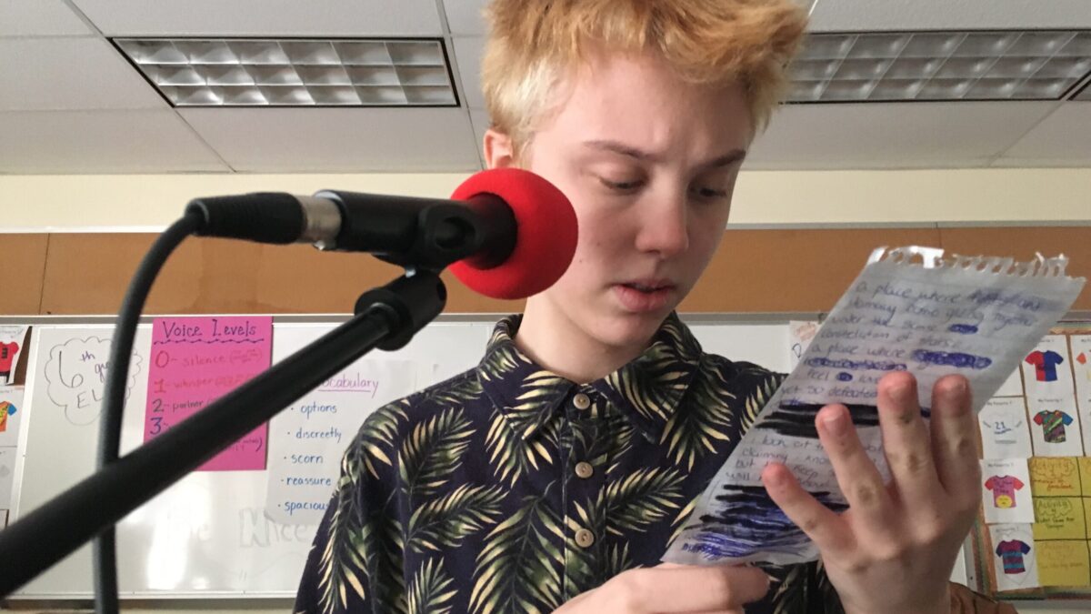 Youth reading from a small piece of notebook paper into a microphone in front of a white board in a classroom