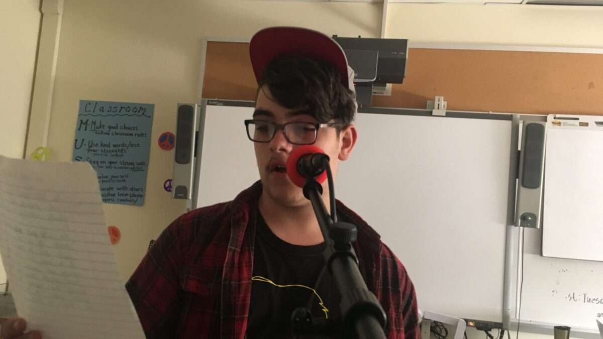Teen looking at a piece of paper and speaking into a microphone in front of a white board in a classroom