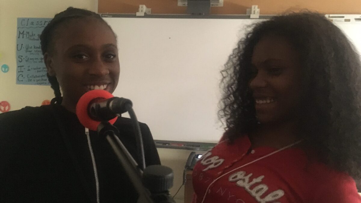 Two teens smiling behind a microphone in front of a white board in a classroom