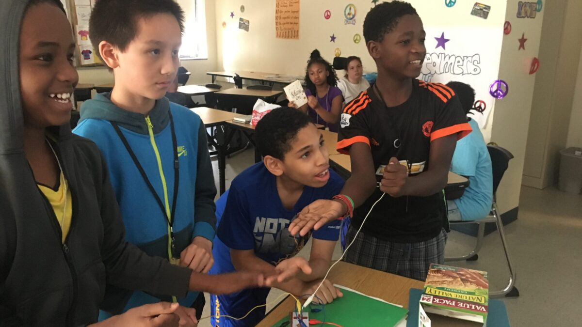 Four youth gathered around a box, three oranges, and wires attached to a controller on a wooden desk