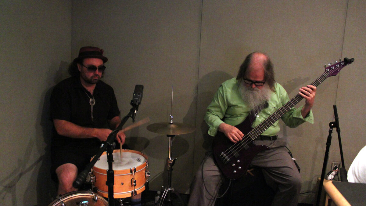 An adult playing drum set and an adult playing electric bass in a radio studio