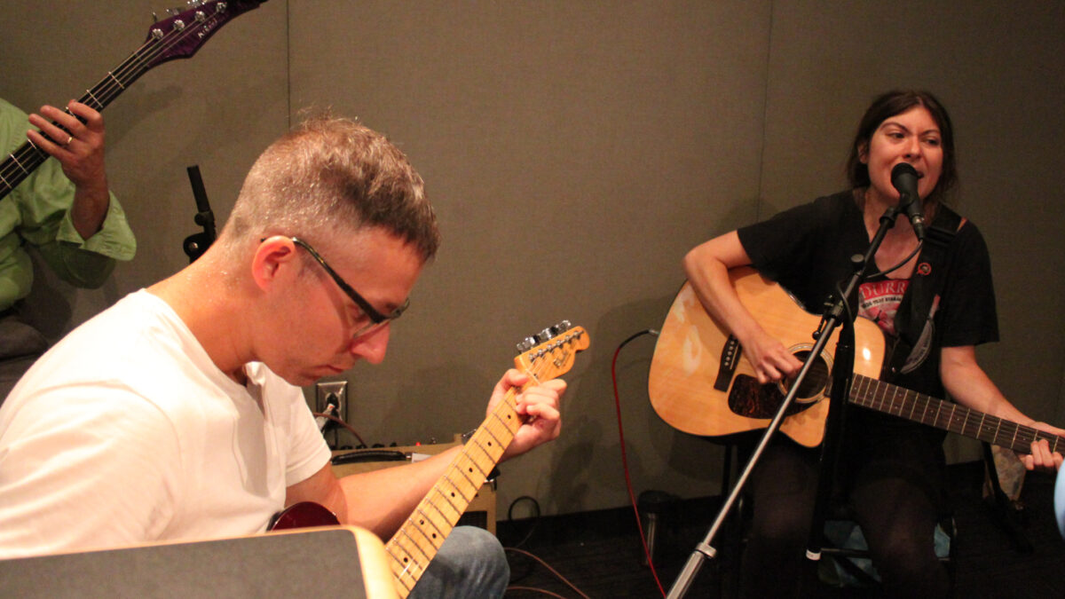 An adult playing electric guitar and an adult playing acoustic guitar and singing into a microphone in a radio studio