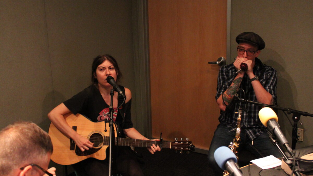 An adult playing acoustic guitar and singing into a microphone and an adult playing harmonica into a microphone in a radio studio