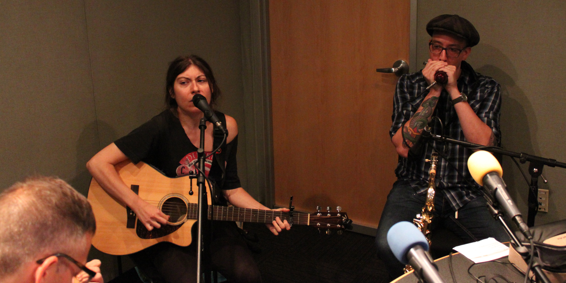 An adult playing acoustic guitar and singing into a microphone and an adult playing harmonica into a microphone in a radio studio