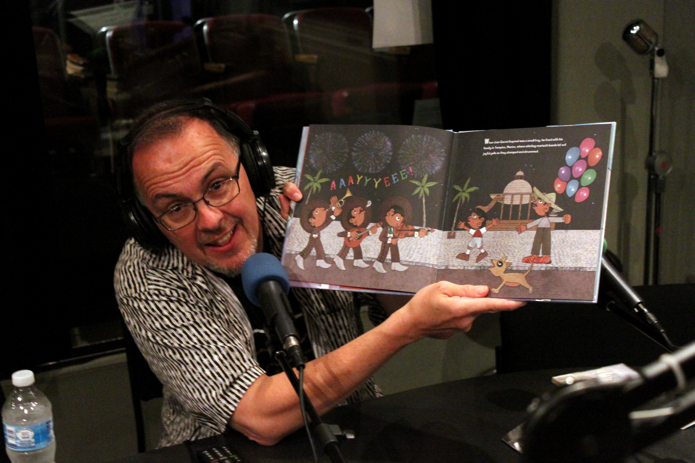 Adult wearing headphones speaking into a microphone and holding an open children's picture book in a radio studio