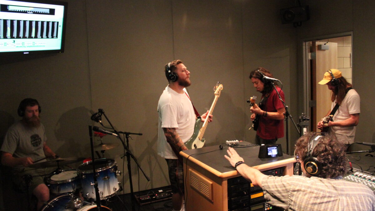An adult playing a drum set, an adult playing electric bass, and two adults playing electric guitar, all wearing headphones, as another adult wearing headphones listens in a radio studio