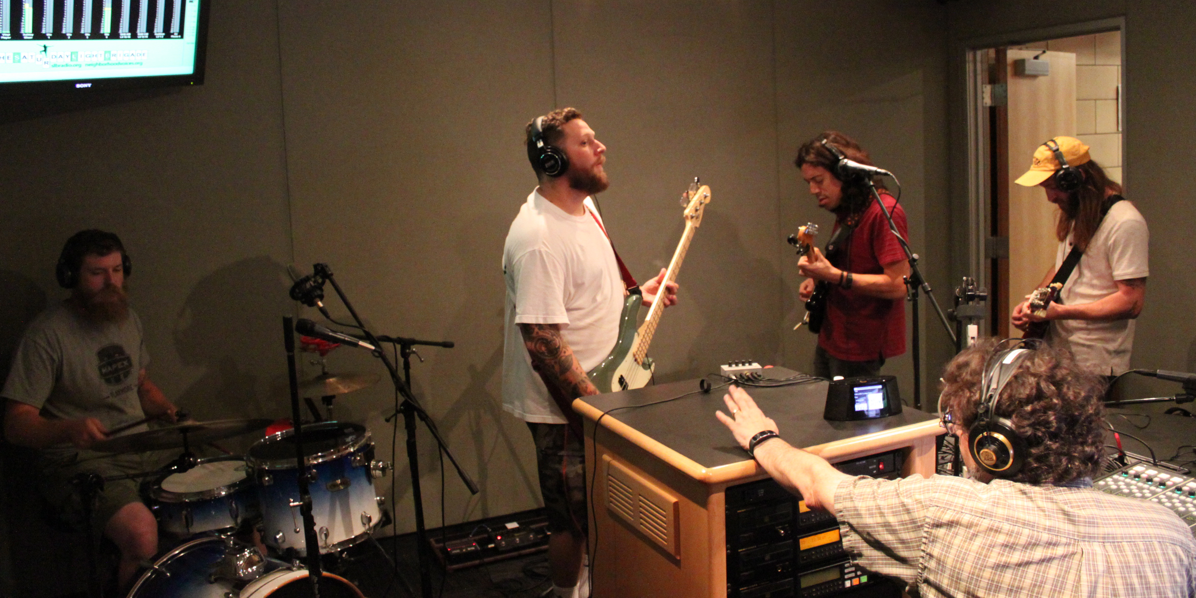 An adult playing drum set, an adult playing electric bass, and two adults playing electric guitar, all wearing headphones as another adult wearing headphones listens in a radio studio