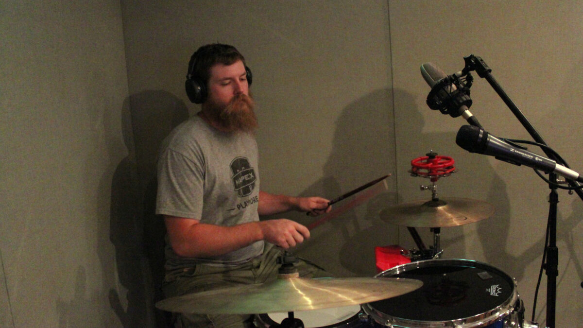 An adult wearing headphones playing a drum set in a radio studio