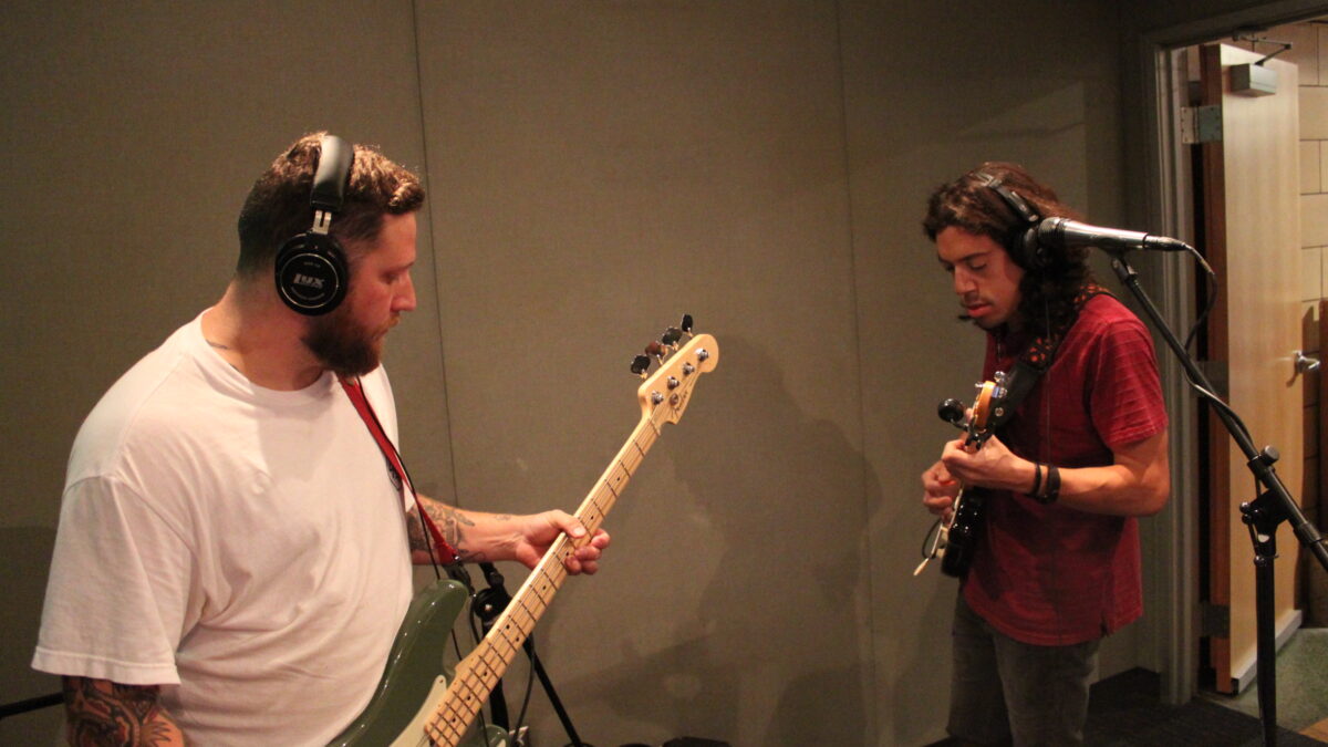 An adult playing electric bass and an adult playing electric guitar, both wearing headphones in a radio studio