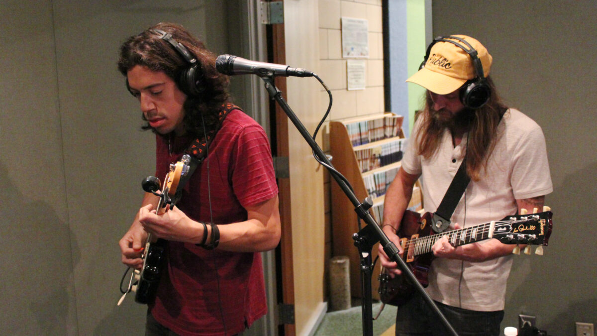 Two adults wearing headphones playing electric guitar in a radio studio