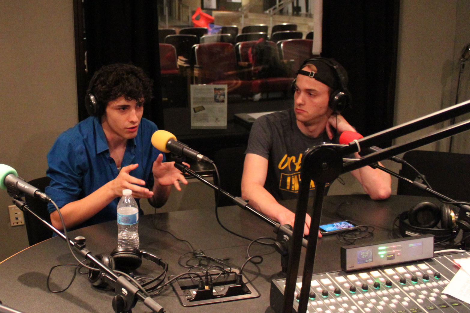 Two teens wearing headphones and speaking into microphones in a radio studio, one gesturing with their hands