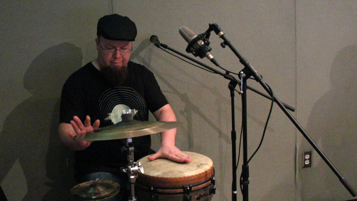 An adult playing a conga and cymbals into microphones in a radio studio