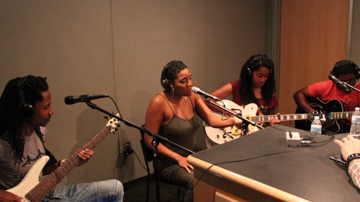 A teen playing electric bass, a teen singing into a microphone, and two teens playing electric guitar, all wearing headphones in a radio studio