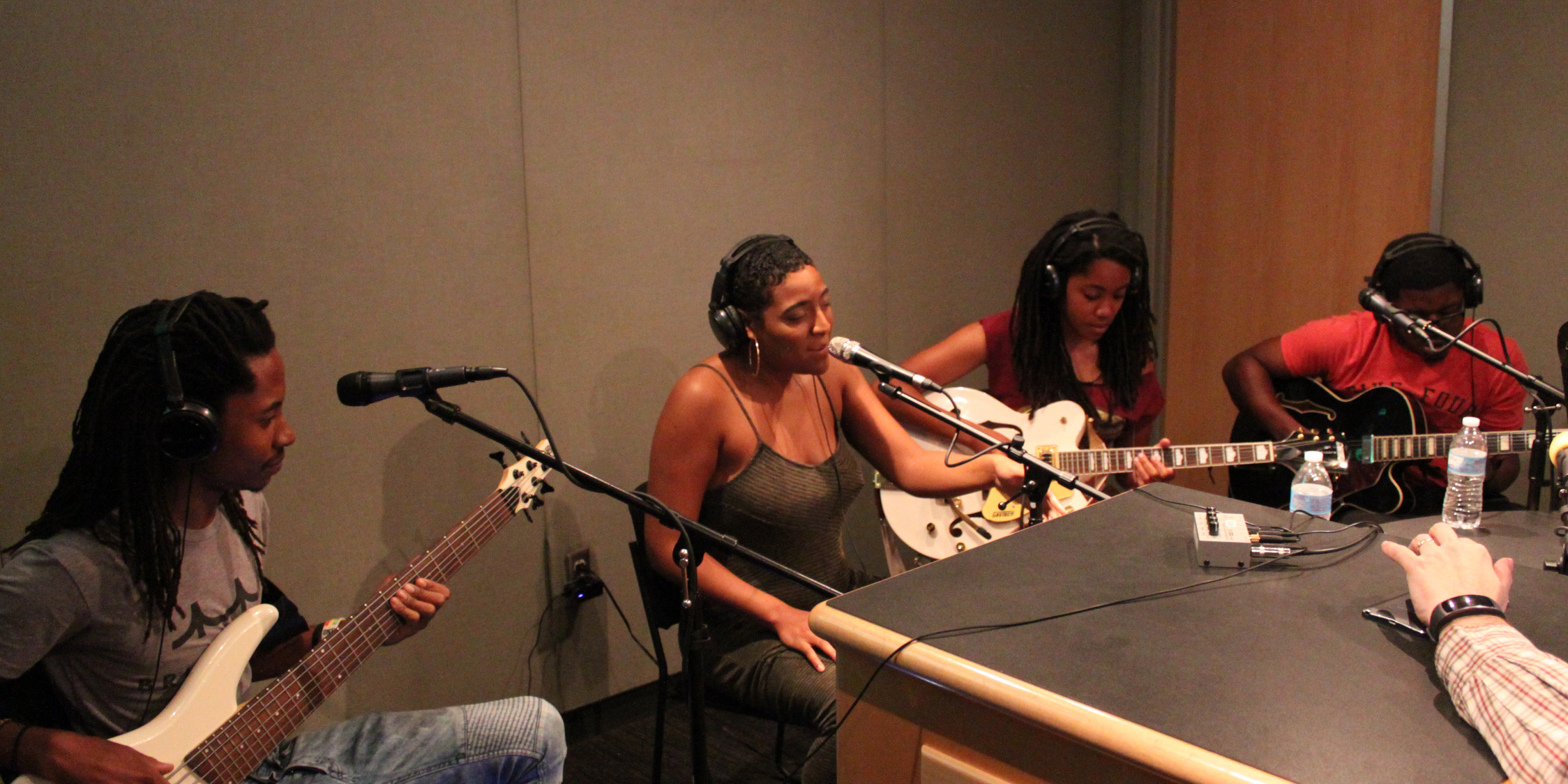 A teen playing electric bass, a teen singing into a microphone, and two teens playing electric guitar, all wearing heaphones in a radio studio