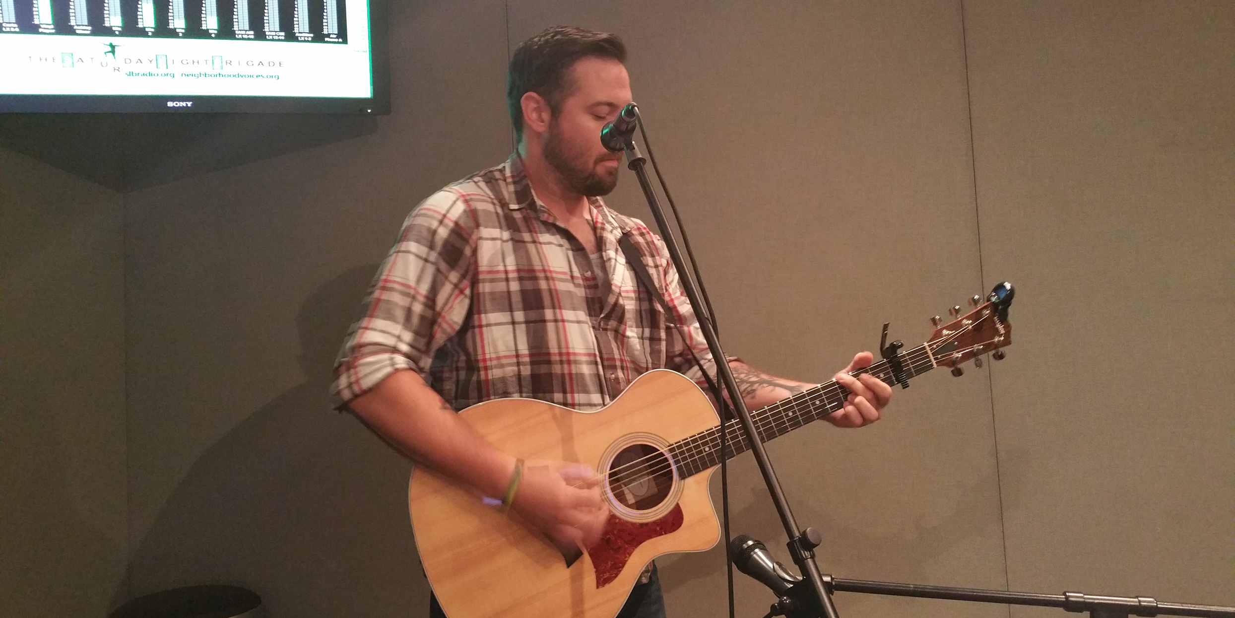 Adult playing acoustic guitar standing behind a microphone in a radio studio