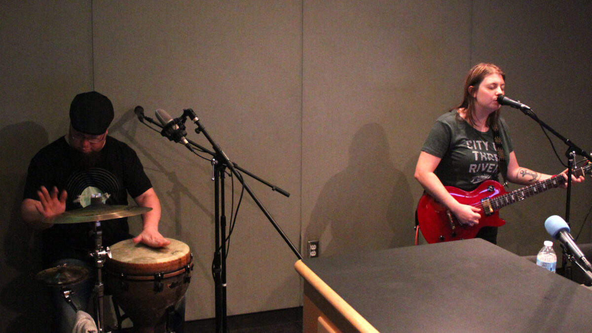 an adult plying a conga and cymbals and an adult playing electric guitar and singing into a microphone in a radio studio