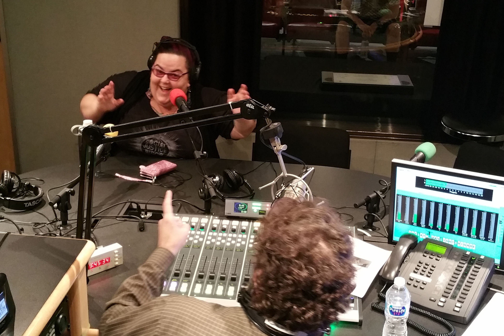 An adult wearing headphones speaking into a microphone and gesturing with their hands being interviewed by an adult wearing headphones sitting behind a microphone and a mixing board in a radio studio