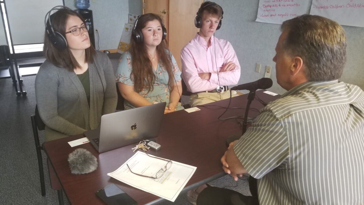 Three teens wearing headphones, one sitting in front of a laptop, interviewing an adult speaking into a microphone on a brown table
