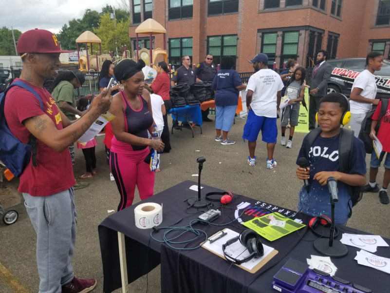 Two adults filming a child wearing headphones and holding a microphone in front of a black table with recording equipment on it, all in front of a small crowd of people in front of a school