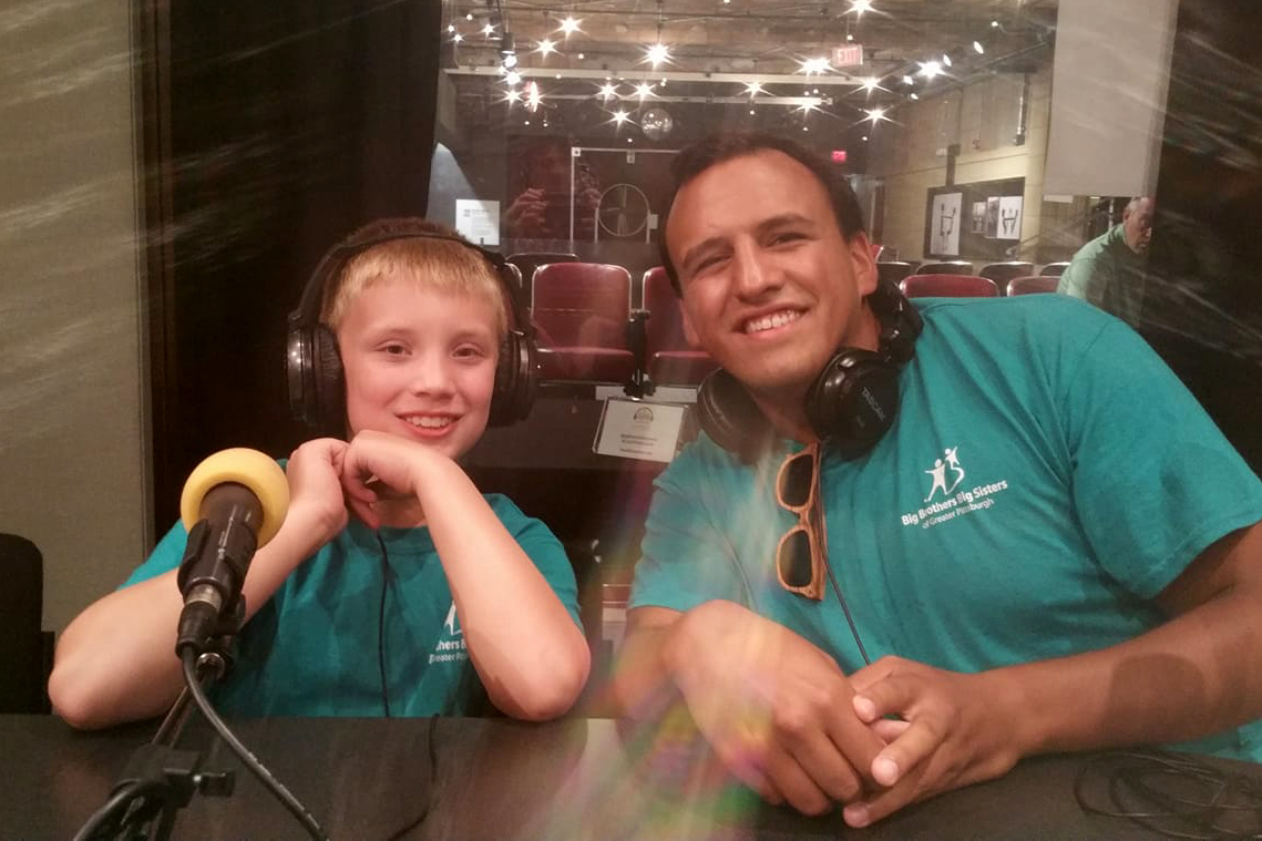 A youth and an adult wearing headphones and sitting behind a microphone smiling in a radio studio