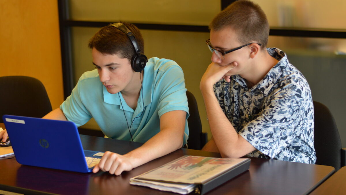 Two teens, one wearing headphones, looking at a laptop on a brown desk