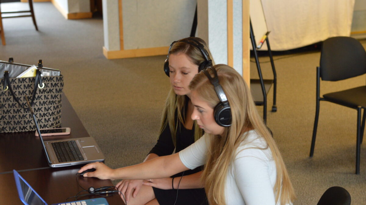 Two teens wearing headphones looking at laptops on brown desks