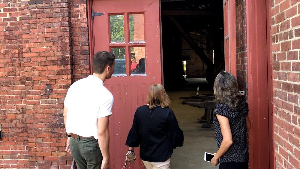 Three individuals heading into a red brick building with a large red door, and a sign above the door reading "Smith Shop"