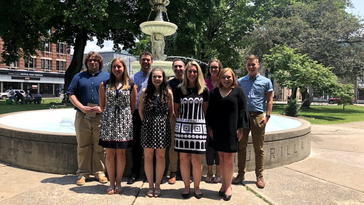 Nine young adults smiling and posing in front of a fountain in a park