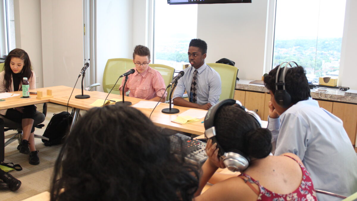 Three teens wearing headphones sitting behind a small mixing board listening to two teens speaking into microphones sitting behind a wooden table