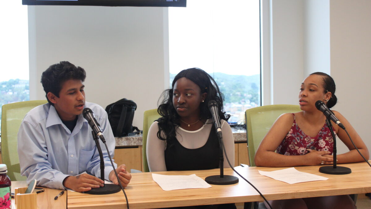 Three teens speaking into microphones sitting behind a wooden table