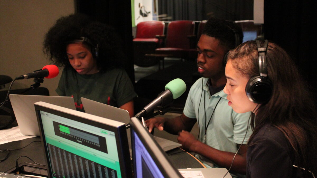Three youth wearing headphones looking at laptops and speaking into microphones in a radio studio