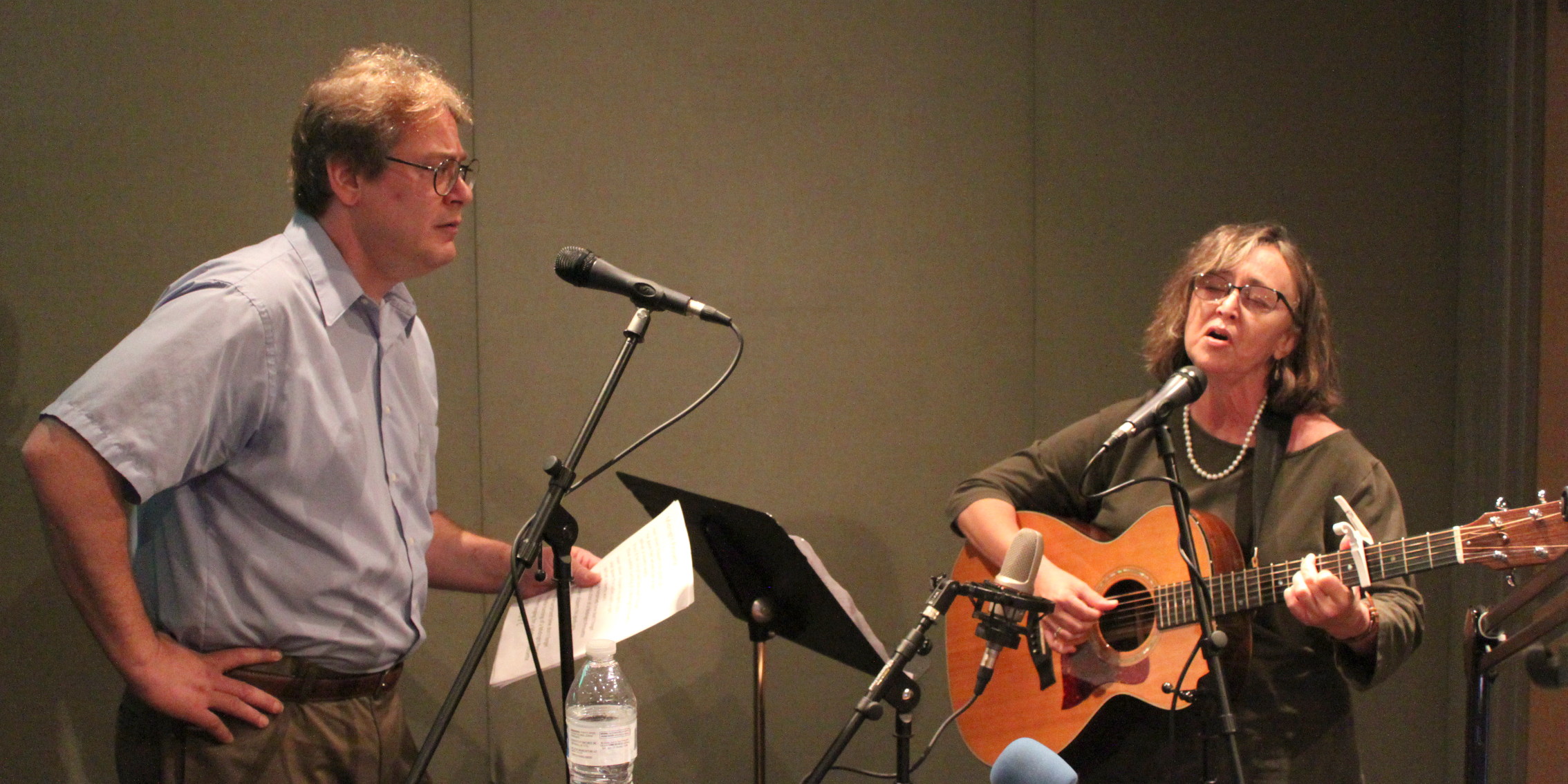 Two adults singing into microphones in a radio studio, one holding a piece of paper, the other playing acoustic guitar