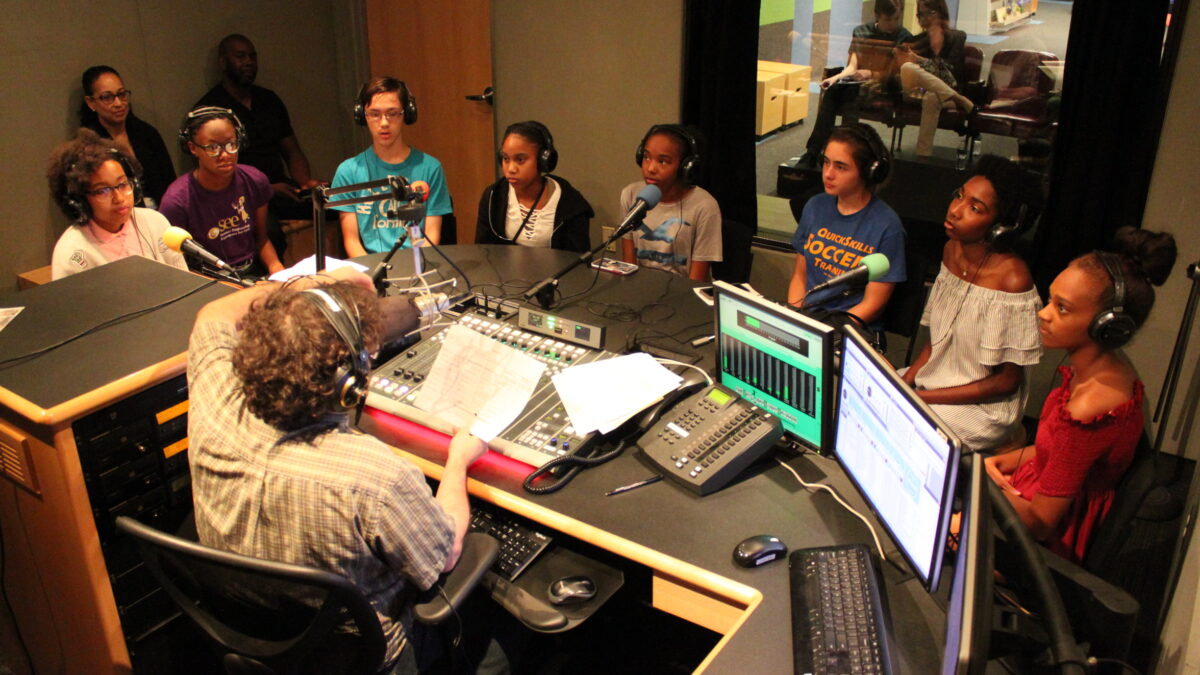 Eight youth wearing headphones speaking into microphones being interviewed by an adult wearing headphones sitting behind a microphone and a mixing board in a radio studio while two adults sit behind the youth listening