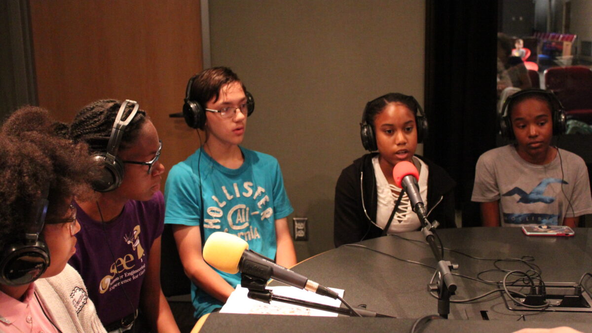Five youth wearing headphones and speaking into microphones in a radio studio