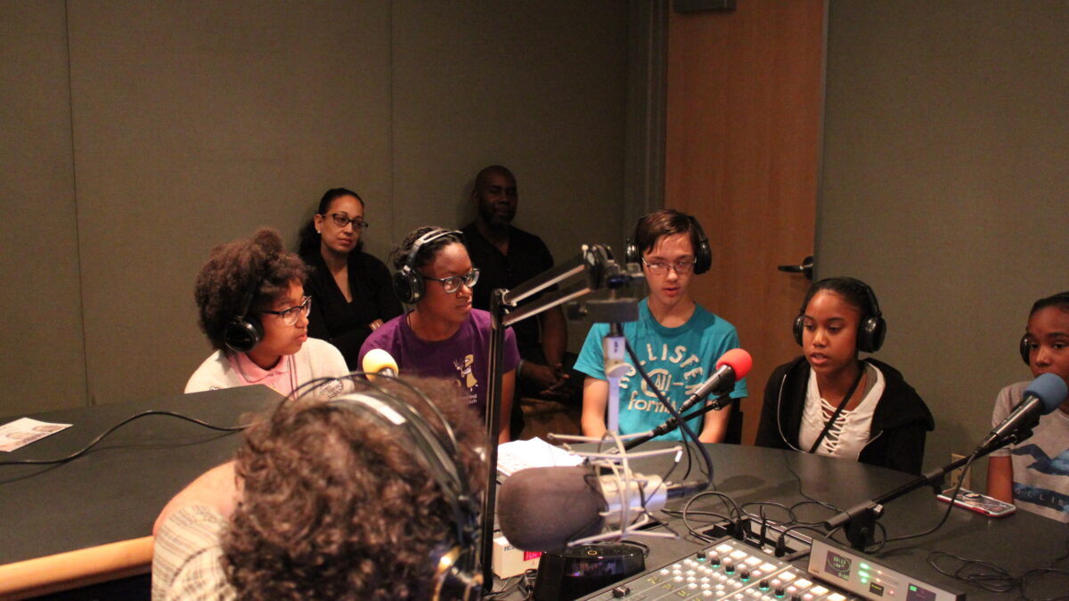 Five youth wearing headphones speaking into microphones being interviewed by an adult wearing headphones sitting behind a mixing board and a microphone in a radio studio while two adults sitting behind the youth listen