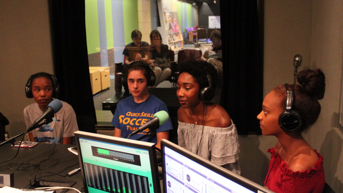 Four teens wearing headphones speaking into microphones in a radio studio