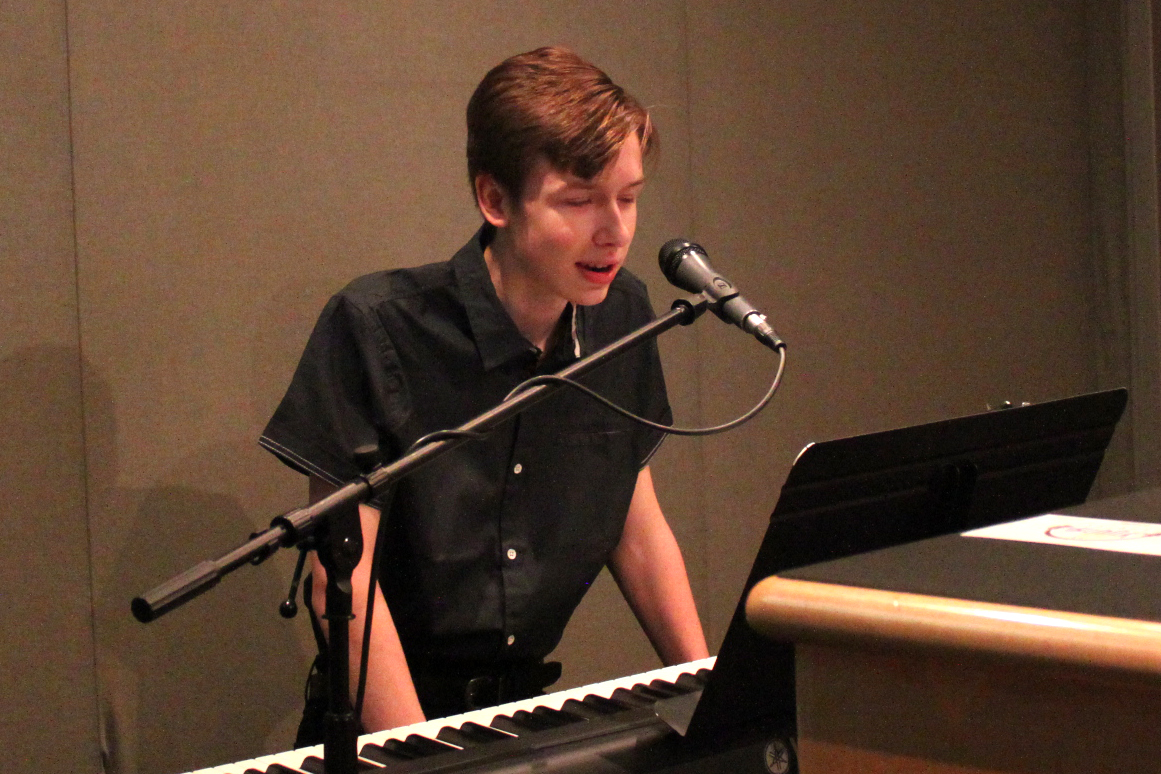 A teen sitting behind a piano keyboard speaking into a microphone in a radio studio