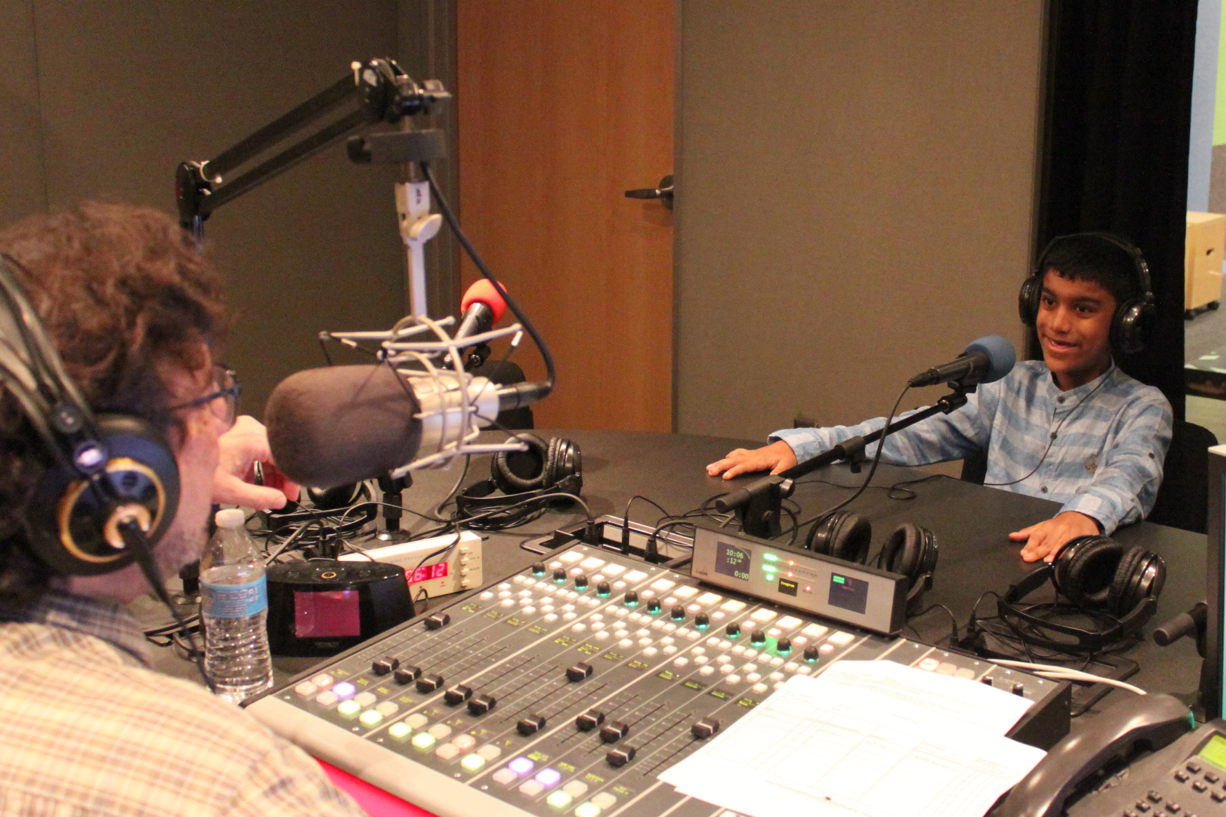 A youth wearing headphones speaking into a microphone being interviewed by an adult wearing headphones sitting behind a microphone and a mixing board in a radio studio