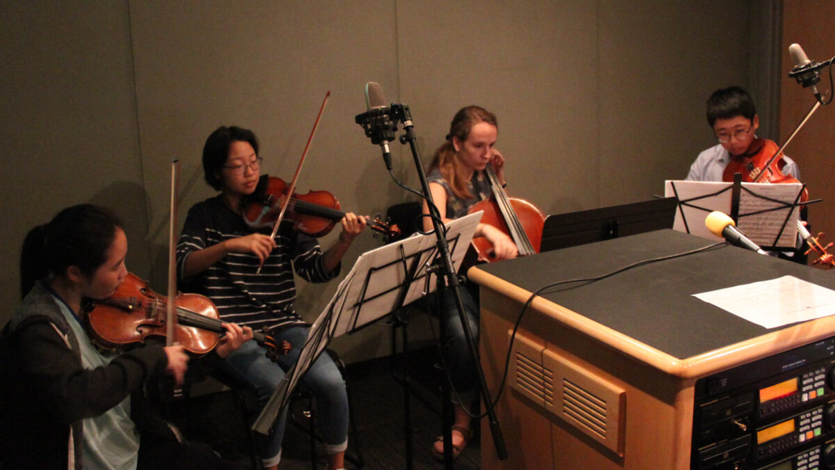 Two teens playing violin, one teen playing cello, and one teen playing viola, all looking at sheet music on music stands in a radio studio