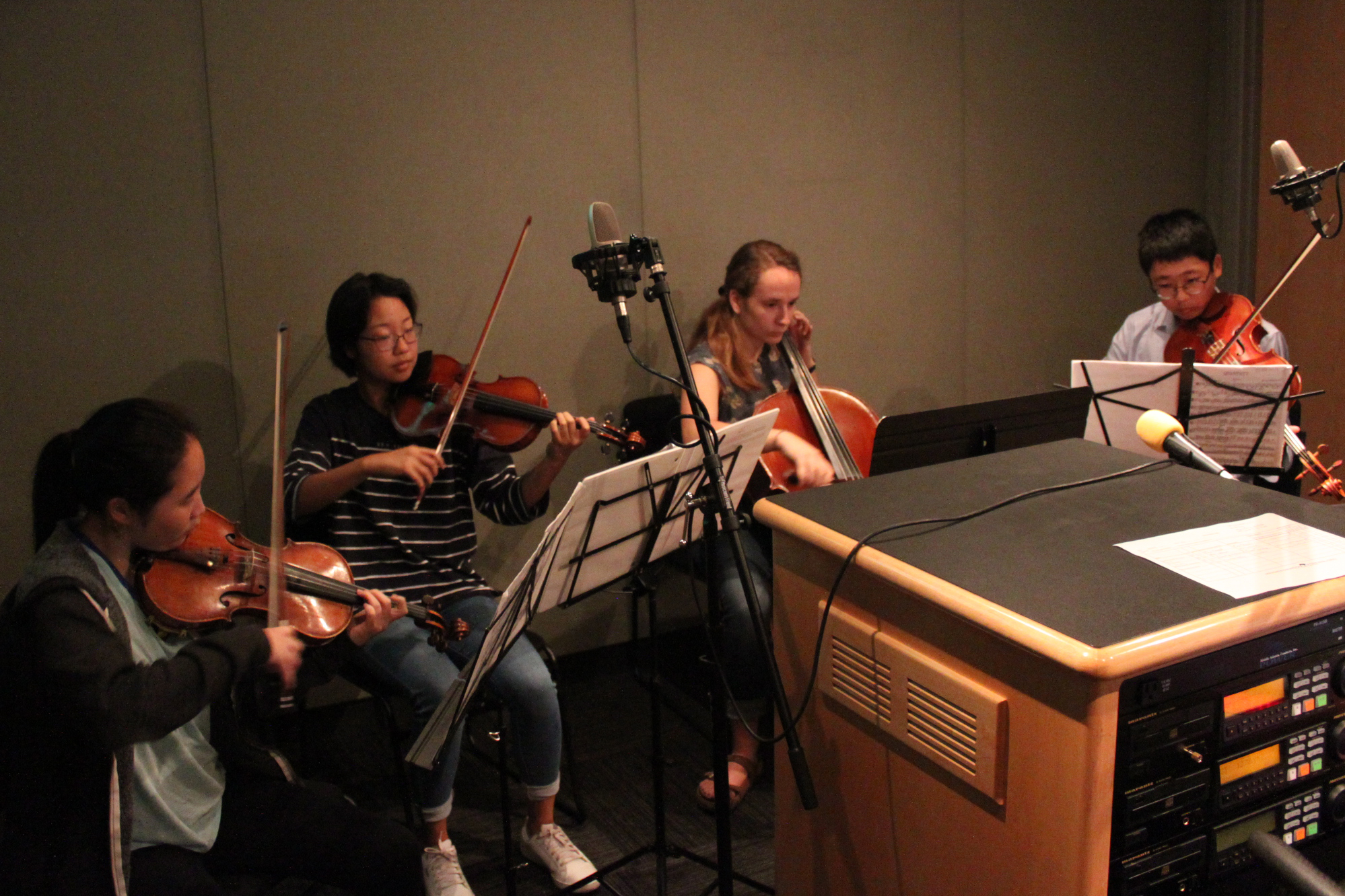 Two teens playing violin, one teen playing cello, and one teen playing viola, all looking at sheet music on music stands in a radio studio