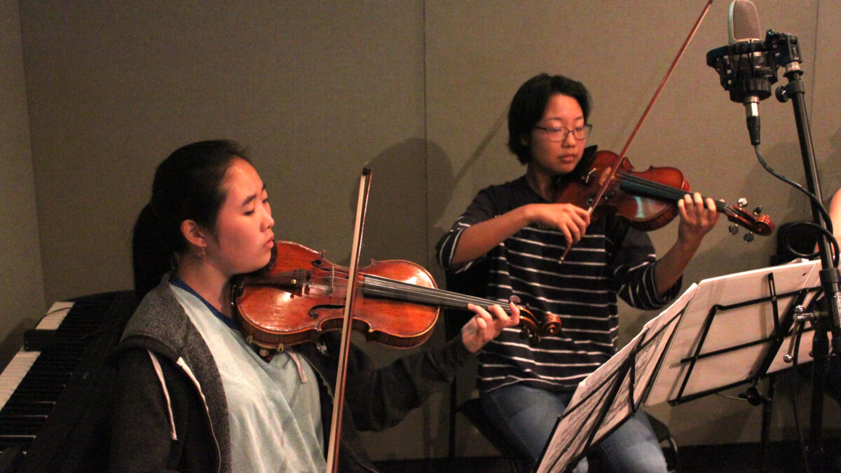 Two youth playing violins and looking at sheet music on music stands in a radio studio