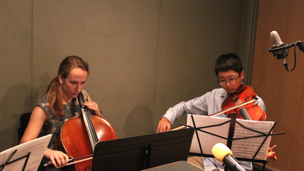 A teen playing cello and a teen playing viola, both looking at sheet music on music stands in a radio studio