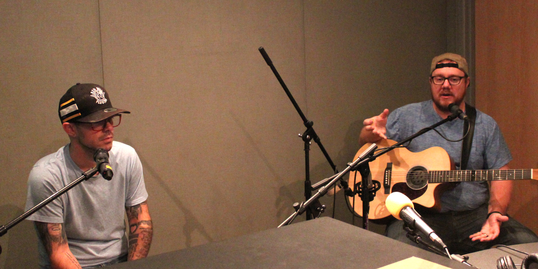 Two adults sitting behind microphones in a radio studio, one holding an acoustic guitar and talking into a microphone