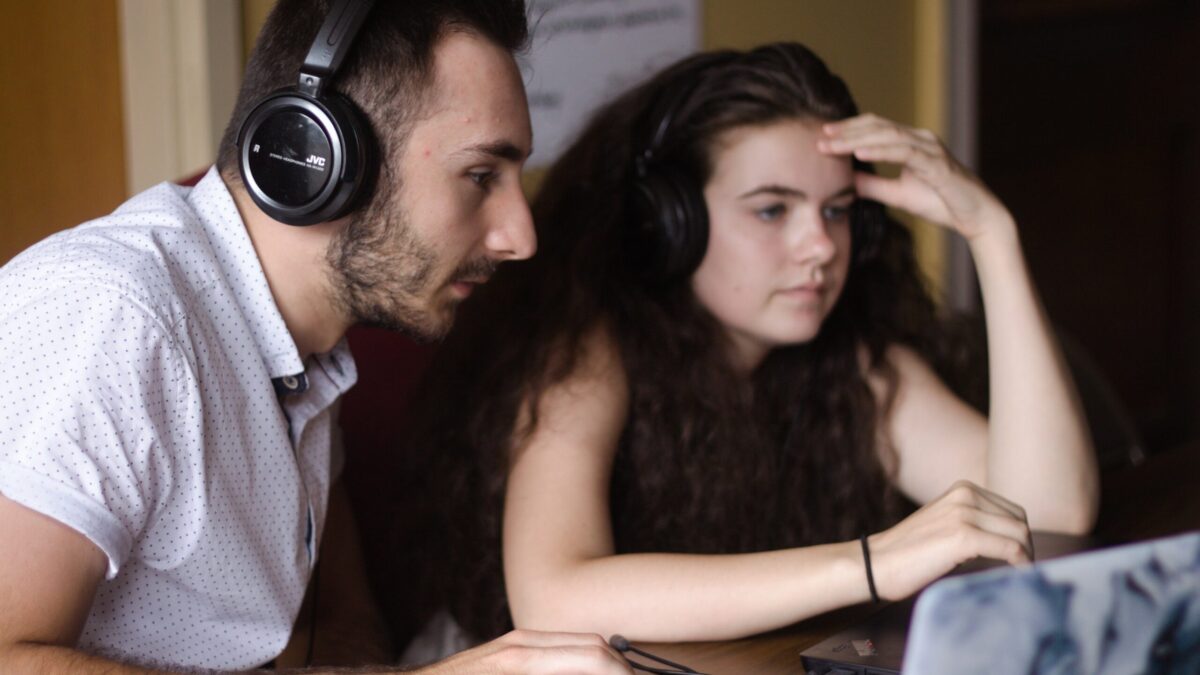 An adult and a teen, both wearing headphones and looking at a laptop